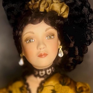 Puede incluir: Una muñeca de porcelana con cabello castaño oscuro, que lleva un tocado de encaje negro y una prenda dorada con patrones florales negros. La muñeca tiene ojos castaños claros, una gargantilla de encaje negro y pendientes colgantes transparentes.