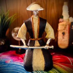Peut inclure: Une figurine de guerrier portant un chapeau à larges bords, un gilet sombre et une robe claire. La figurine a une tête et des mains argentées brillantes et tient une longue épée argentée horizontale devant le corps.