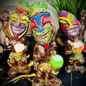 May include: Three Tiki statues with smiling faces and a wood-like texture. Each statue features a unique colour scheme, including green, blue, red, and yellow. One holds a green coconut, another a mug of beer, and the third a white object.