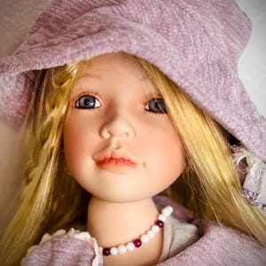 May include: A porcelain doll with blonde hair and blue eyes, wearing a lilac hat and matching coat. The doll has a delicate face with painted features and a beaded necklace. The doll's outfit has white lace trim.