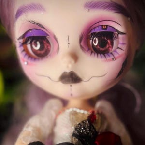 Puede incluir: Una muñeca de piel pálida, cabello morado y grandes ojos marrones con sombra de ojos morada y pestañas oscuras. El rostro tiene labios negros pintados, una línea fina que sale de la boca y pequeños brillantes cerca de los ojos. La muñeca viste un conjunto blanco y negro con detalles de encaje y un acento rojo.