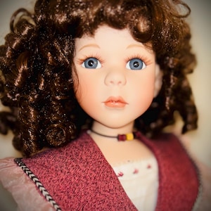 May include: A porcelain doll with curly brown hair, blue eyes, and long eyelashes. The doll wears a textured maroon vest over a cream-colored blouse with small red floral accents. A thin black cord necklace with two small square beads rests on the neck.