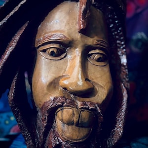 May include: A hand-carved wooden sculpture of a human head featuring detailed facial features, including a moustache and beard. The sculpture displays a smooth, polished finish on the face with textured, dark brown carved hair or dreadlocks.