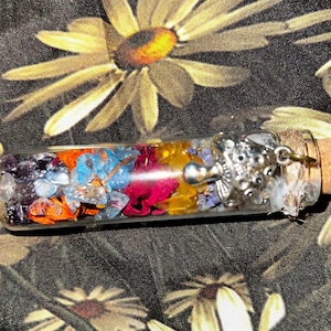 Puede incluir: Un vial de vidrio transparente lleno de piedras preciosas de colores y flores secas. El vial tiene un tapón de metal plateado y una tapa de corcho. El contenido incluye elementos morados, azules, naranjas, rojos y amarillos. El vial mide aproximadamente 10 cm de largo.