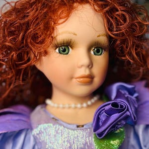 Może przedstawiać: Porcelanowa lalka z rudymi kręconymi włosami i zielonymi oczami. Lalka ma na sobie fioletową sukienkę z perłowym naszyjnikiem i materiałową różą. Sukienka ma cekinowy gorset. Klasyczna lalka kolekcjonerska.