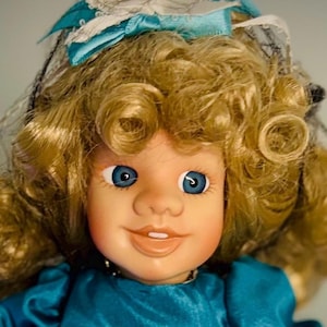 Może przedstawiać: Porcelanowa lalka z blond, kręconymi włosami i jasnoniebieskimi oczami. Lalka ma na sobie turkusową sukienkę i pasujący czepek z niebieską kokardą. Lalka ma uśmiechnięty wyraz twarzy.