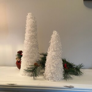 May include: Two white fluffy cone-shaped Christmas trees on a white distressed wood surface with a small green garland with red berries.