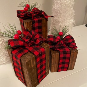 May include: Three wooden boxes wrapped in red and black buffalo plaid ribbon with a red berry sprig on top. The boxes are arranged in a pyramid shape on a white surface.