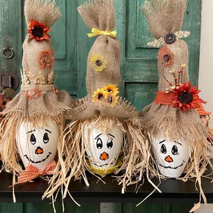 May include: Three whimsical scarecrow decorations. Each has a white painted head with a smiling face, straw hair, and a burlap body. They are adorned with sunflowers, ribbons, and other fall-themed embellishments. The scarecrows are displayed on a black shelf.