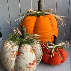 May include: Three fabric pumpkins in different sizes. The largest pumpkin is orange, the middle pumpkin is beige with a fall leaf pattern, and the smallest pumpkin is orange. All pumpkins have a brown twig stem and are decorated with straw and green moss.