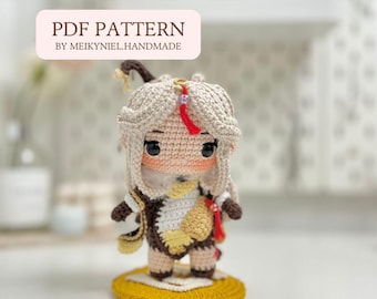 May include: Crochet amigurumi doll pattern PDF. The doll is a light brown color with a white and yellow outfit. The doll has long blonde hair and a red ribbon. The doll is standing on a yellow crocheted base. The text "PDF PATTERN BY MEIKYNIEL.HANDMADE" is on a pink rectangle.