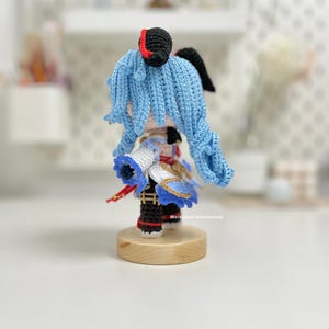 May include: A crocheted blue-haired character with a red and black hat, wearing a white and blue outfit with gold accents. The character is standing on a wooden base.