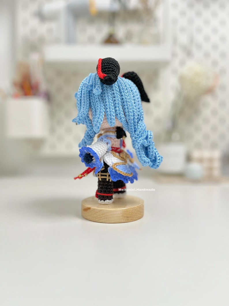 May include: A crocheted blue-haired character with a red and black hat, wearing a white and blue outfit with gold accents. The character is standing on a wooden base.