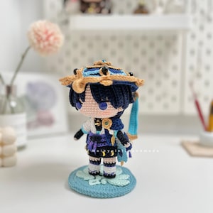 Op de afbeelding: Gehaakte figuur van een personage met blauw haar, met een blauwe en gouden hoed, een blauwe en paarse outfit en staand op een blauwe en groene basis.