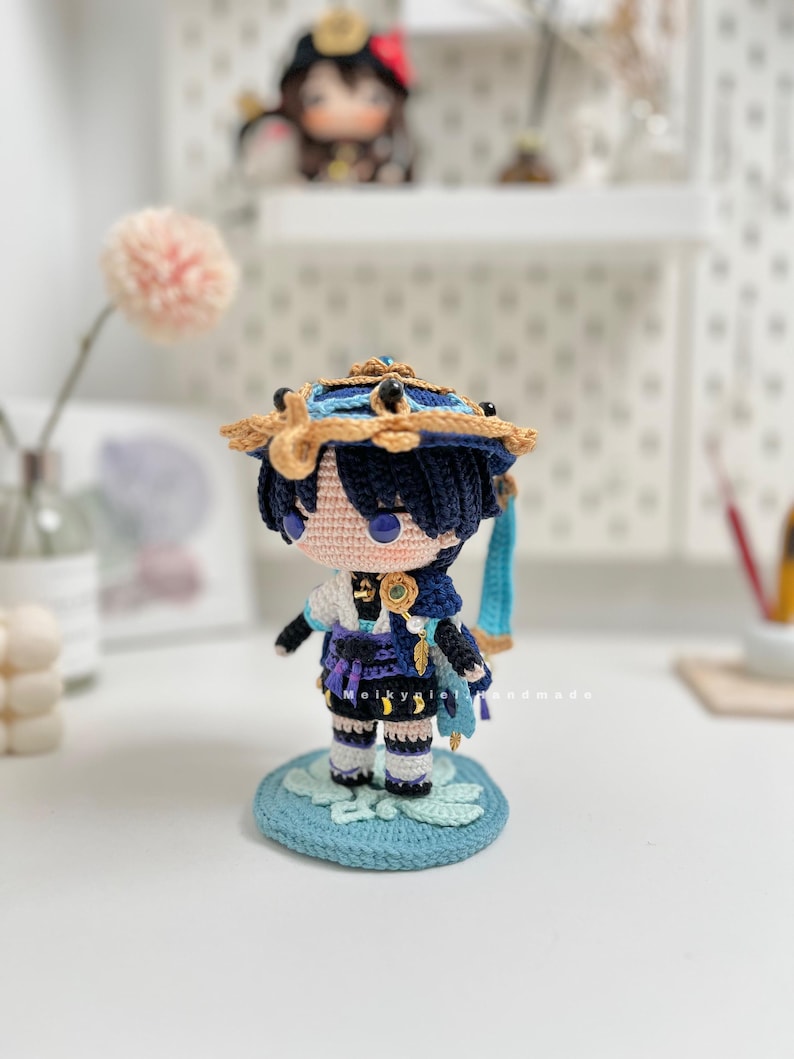 Op de afbeelding: Gehaakte figuur van een personage met blauw haar, met een blauwe en gouden hoed, een blauwe en paarse outfit en staand op een blauwe en groene basis.