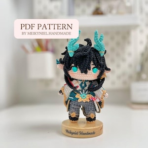 以下が含まれることがあります： 黒髪にターコイズブルーの角を持つキャラクターの、かぎ針編みアミグルミ人形。ターコイズブルーとゴールドの衣装を着ており、"Meikyniel Handmade"という文字が書かれた木製の台座の上に立っている。