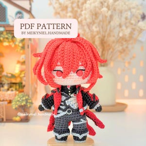 Puede incluir: Una muñeca amigurumi de ganchillo con cabello rojo brillante, tez pálida y un atuendo detallado en negro y gris con acentos rojos. La muñeca tiene un pequeño detalle de joya roja en el pecho. Una etiqueta arriba dice PDF PATTERN BY MEIKYNIEL.HANDMADE.
