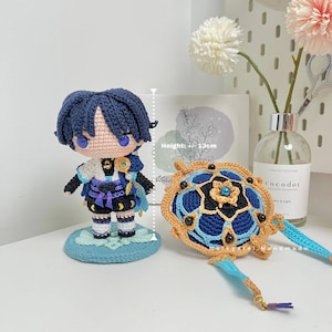 Op de afbeelding: Gehaakte blauwe en gouden figuur met zwart haar en blauwe ogen, staand op een blauwe basis. De figuur is ongeveer 12 cm hoog. Een gehaakt blauw en gouden mandala met een blauwe en gouden kwast is ook afgebeeld.
