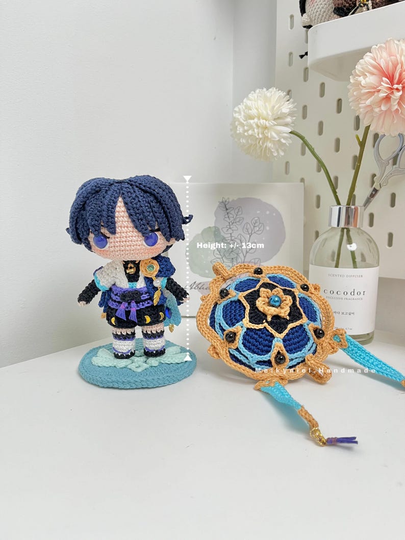 Op de afbeelding: Gehaakte blauwe en gouden figuur met zwart haar en blauwe ogen, staand op een blauwe basis. De figuur is ongeveer 12 cm hoog. Een gehaakt blauw en gouden mandala met een blauwe en gouden kwast is ook afgebeeld.