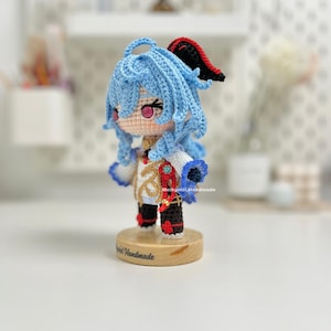 May include: A crocheted doll with blue hair, a white and gold outfit, and red shoes. The doll is standing on a wooden base with the text "Melkyniel Handmade" on it.