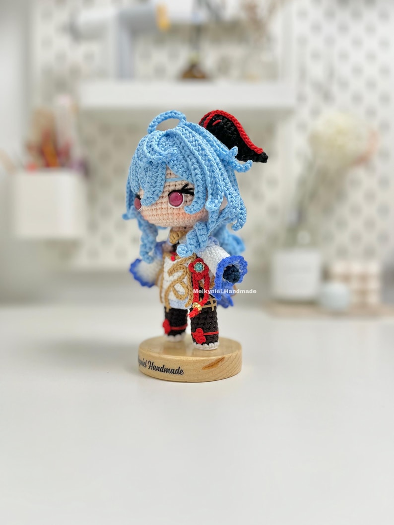 May include: A crocheted doll with blue hair, a white and gold outfit, and red shoes. The doll is standing on a wooden base with the text "Melkyniel Handmade" on it.