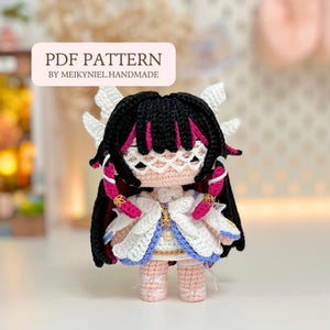 May include: A handmade crochet doll with black hair, pink highlights, and white wings. The doll wears a white dress with blue trim and gold accents. The text "PDF PATTERN BY MEIKYNIEL.HANDMADE" is displayed on a white banner.