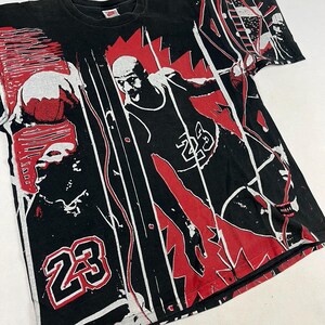 May include: Black t-shirt featuring a graphic design of a basketball player in action, with the number 23 prominently displayed. The design incorporates red and white accents, creating a dynamic and bold visual.