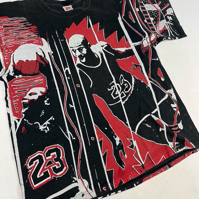 May include: Black t-shirt featuring a graphic design of a basketball player in action, with the number 23 prominently displayed. The design incorporates red and white accents, creating a dynamic and bold visual.
