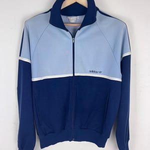 Può includere: Una giacca sportiva bicolore con una sezione superiore azzurra e una sezione inferiore blu scuro, separate da una sottile striscia orizzontale bianca. La giacca presenta un colletto alto, una cerniera frontale e il logo adidas sul petto.