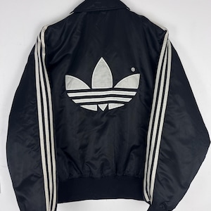 Może przedstawiać: Czarna kurtka typu bomber z dużym, białym, wyhaftowanym logo koniczyny Adidas na plecach. Kurtka posiada klasyczny kołnierz, długie rękawy z trzema białymi paskami wzdłuż każdego ramienia oraz czarne ściągacze u dołu i przy mankietach.