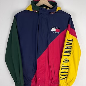 May include: A color-blocked hooded jacket featuring navy blue, red, green, and yellow panels. The chest displays a Tommy Jeans logo patch, while the left sleeve features the text Tommy Jeans in black lettering with a circular emblem.