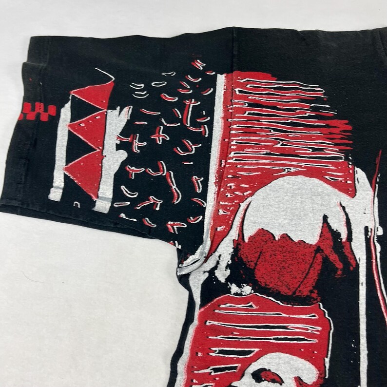 May include: Black t-shirt with a graphic design in red and white. The design features abstract shapes and lines, with a central figure. The shirt's sleeve is visible, showing the fabric's texture.
