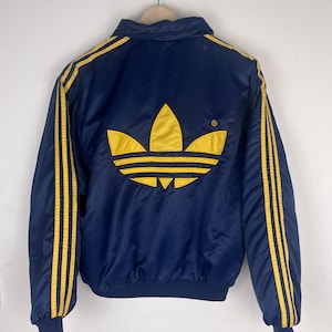 May include: A navy blue satin jacket featuring a large yellow embroidered trefoil logo on the back. The jacket has long sleeves with three yellow stripes running down each arm, a ribbed collar, and ribbed cuffs at the wrists and waist hem.