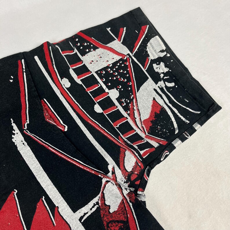 May include: Black t-shirt with a graphic design featuring red and white abstract shapes. The design includes lines, rectangles, and splattered paint effects. The shirt is made of a soft, likely cotton material.