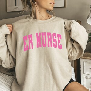 May include: A light beige sweatshirt with the text "ER NURSE" in pink lettering.