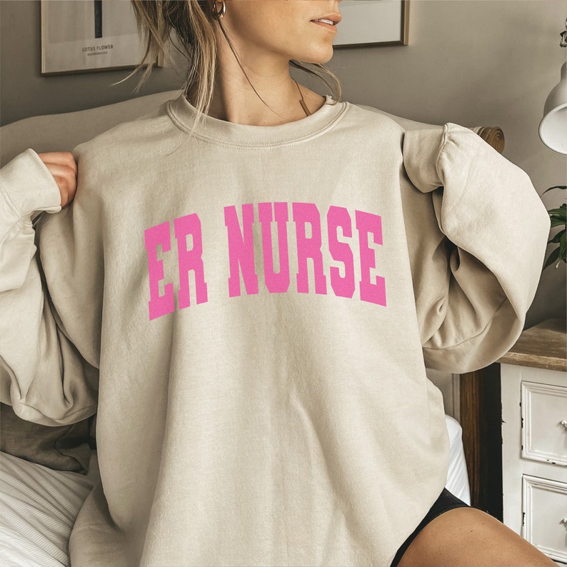 May include: A light beige sweatshirt with the text "ER NURSE" in pink lettering.