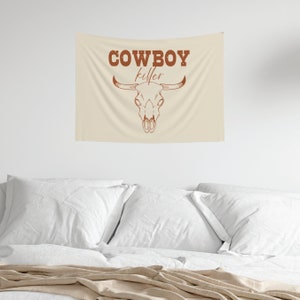 May include: A beige tapestry with the text "COWBOY killer" in brown lettering. A brown bull skull with horns is centered below the text.