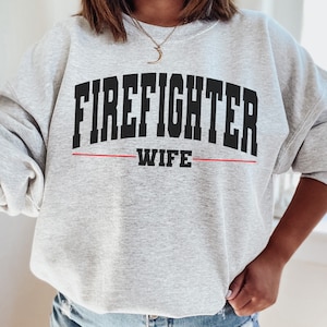 May include: Grey sweatshirt with the text "FIREFIGHTER WIFE" in black letters with a red line underneath.