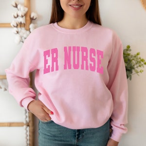 May include: Pink sweatshirt with the text "ER NURSE" in a large, white font.