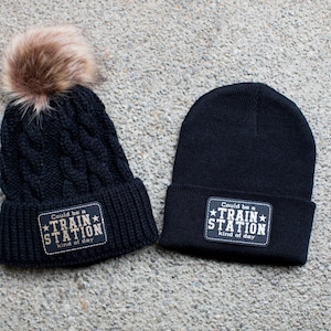 May include: Two black knit beanies with pom poms. One beanie has a gold patch that says "Could be a *TRAIN* STATION kind of day". The other beanie has a similar patch that says "Could be a *TRAIN* STATION kind of day".
