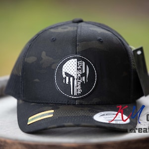 May include: Black and green camouflage trucker hat with a white skull and American flag design. The hat has a black leather patch with the text "We The People" in white.
