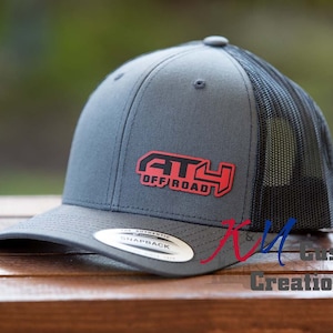 May include: Gray and black trucker hat with the text "ATV OFF ROAD" in red and black. The hat has a curved brim and a mesh back. The hat is sitting on a wooden surface.