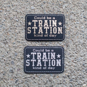 May include: Two black leather patches with white stitching. The patches have silver text that reads "Could be a *TRAIN* STATION kind of day".