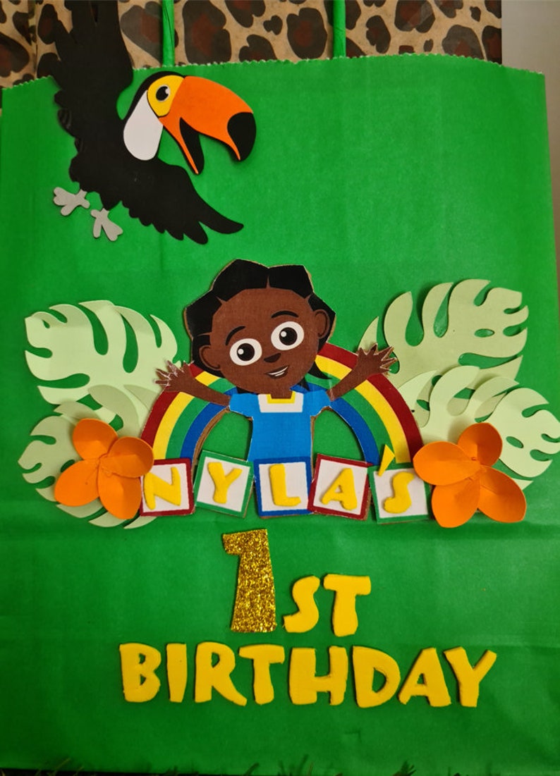 May include: Green gift bag with a rainbow, a cartoon character, and the words "NYLA'S 1ST BIRTHDAY" in yellow letters.