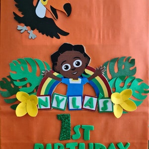 May include: Orange paper gift bag with a jungle theme. It features a toucan, a girl with a rainbow, green leaves, yellow flowers, and the words "Nyla's 1st Birthday" in green letters.
