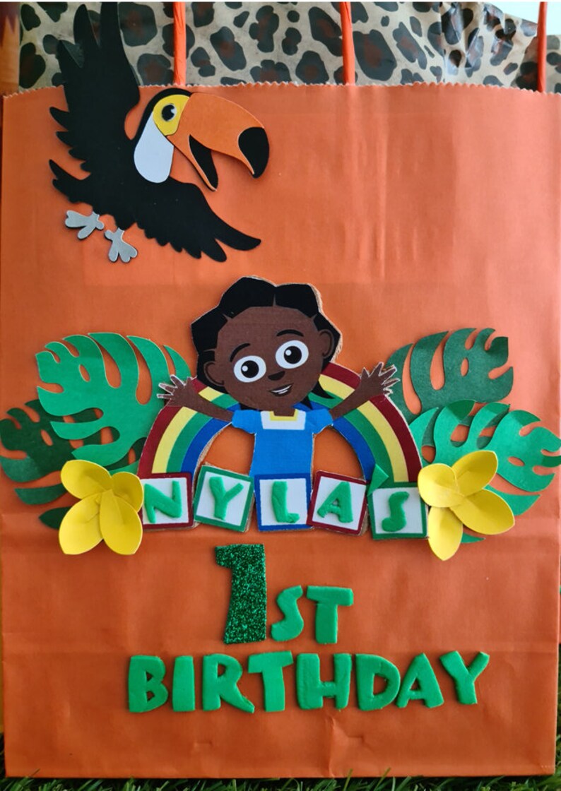 May include: Orange paper gift bag with a jungle theme. It features a toucan, a girl with a rainbow, green leaves, yellow flowers, and the words "Nyla's 1st Birthday" in green letters.