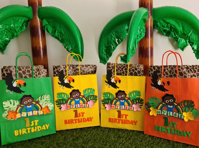 May include: Four green, yellow, and orange paper gift bags with jungle animal themes. Each bag has a toucan, a brown-skinned child, and the text "1st Birthday" and "NYLAS".