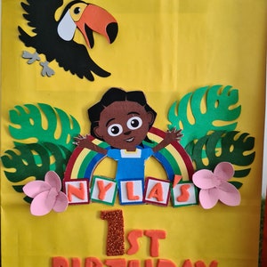 May include: A yellow paper gift bag decorated with a toucan, a girl with a rainbow, green leaves, pink flowers, and the text "NYLA'S 1st BIRTHDAY".