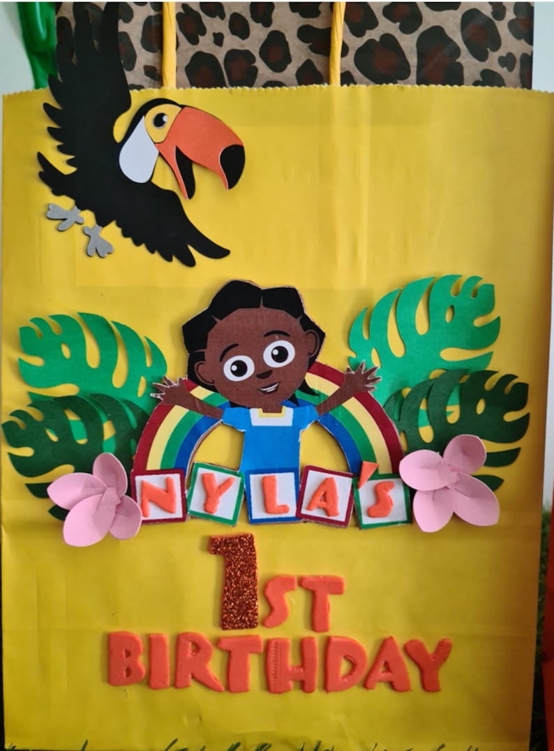 May include: A yellow paper gift bag decorated with a toucan, a girl with a rainbow, green leaves, pink flowers, and the text "NYLA'S 1st BIRTHDAY".