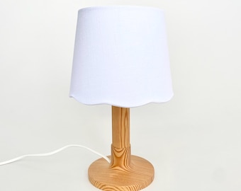 May include: A small table lamp with a white scalloped lampshade. The lamp has a light-colored wooden base and stem. A white electrical cord extends from the base.